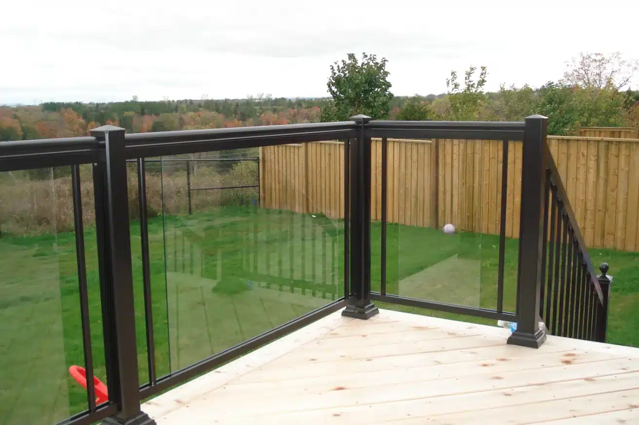 A light-colored wood deck with a black aluminum railing system featuring large glass panels, overlooking a green lawn with a wooden privacy fence in the background.