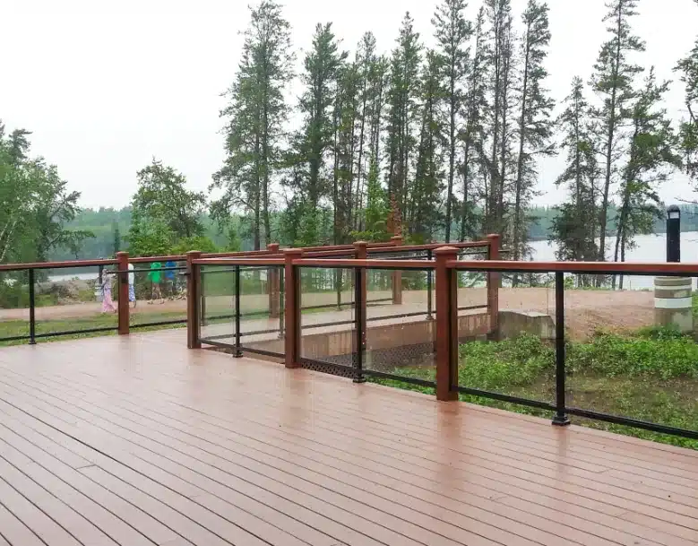 A wooden deck featuring a railing system with thin black aluminum frames and clear glass panels, overlooking a lake and forest.