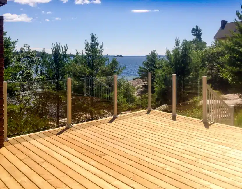 Modern wood deck with topless glass railings overlooking a lake and trees.