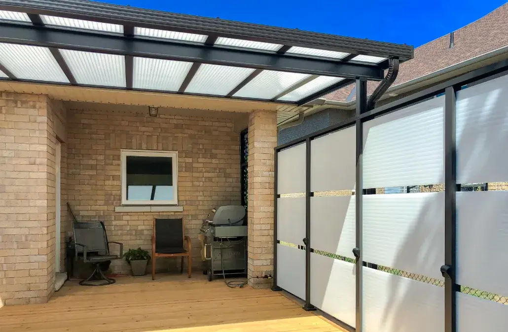 Black metal pergola with a white translucent roof and privacy wall over a wooden deck with grey wicker furniture.