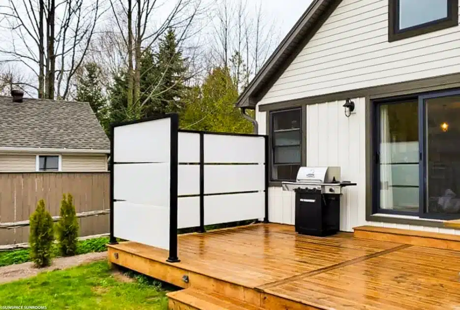 A wooden deck featuring a frosted white horizontal slat privacy screen with a black aluminum frame next to a BBQ grill.